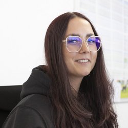 Eine Frau mit langen, braunen Haaren und einer schwarzen Kapuzenjacke lächelt freundlich in die Kamera.