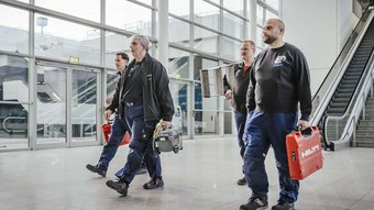 Die Gruppe von Technikern trägt verschiedene Werkzeuge und geht zielstrebig in einem modernen Gebäude mit Treppen und Aufzügen.