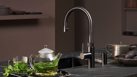 Ein eleganter Wasserhahn steht neben einer Teekanne und einem Glas mit frischem Minztee auf einer dunklen Arbeitsplatte.