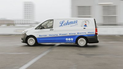 Ein Lieferwagen mit dem Logo von Lohmar fährt über einen Parkplatz, umgeben von modernen Gebäuden und grauem Himmel.