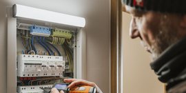 Ein Mann mit Bart überprüft eine elektrische Schalttafel mit einem Messgerät in der Hand, konzentriert auf die Arbeit.
