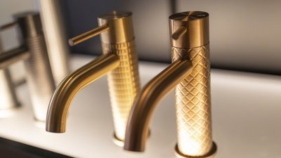 Zwei elegante Wasserhähne aus gebürstetem Gold stehen nebeneinander auf einer hellen Oberfläche, beleuchtet von sanftem Licht.