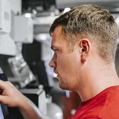 Ein Mann in einem roten T-Shirt berührt einen Bildschirm, während er in einem modernen technischen Raum arbeitet.