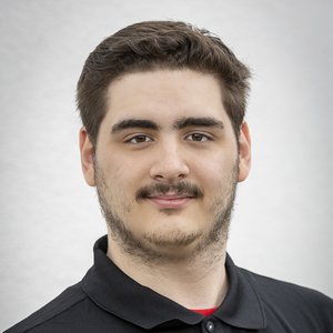 Der Mann hat dunkles, kurzes Haar und schaut direkt in die Kamera, während er vor einem hellen Hintergrund steht.