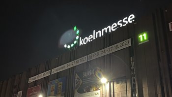 Die beleuchtete Fassade der Koelnmesse zeigt das Logo und die Nummer 11, umgeben von Werbeplakaten und Nachtlicht.