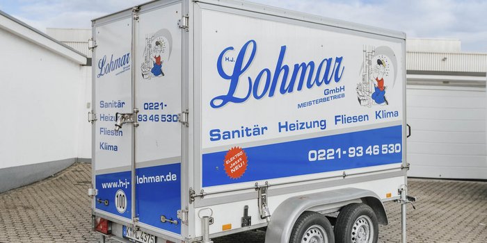 Der Anhänger von Lohmar GmbH zeigt eine bunte Werbung für Sanitär- und Heizungsdienstleistungen, umgeben von einem modernen Gebäude.