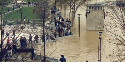 Eine Gruppe von Menschen steht an einem überfluteten Ufer, während Wasser die Umgebung umschließt und Bäume teilweise unter Wasser stehen.