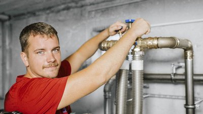Ein junger Mann mit blauen Augen und einem Schnurrbart arbeitet konzentriert an einer Rohrleitung in einem grauen Raum.