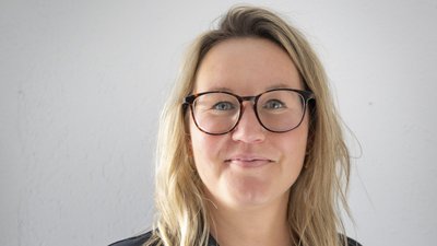 Die Frau mit Brille hat einen freundlichen Ausdruck und trägt ein schwarzes Oberteil, während sie in die Kamera schaut.