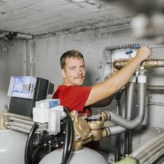 Ein junger Mann in einem roten Shirt arbeitet an Rohren und Ventilen in einem technischen Raum mit Maschinen.