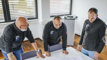 Die Männer stehen um einen Tisch herum und betrachten aufmerksam ein Blatt Papier, das auf dem Tisch ausgebreitet ist.