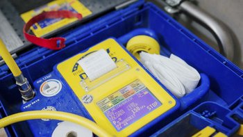 Ein blauer Kasten enthält ein digitales Messgerät mit einem gelben Display und einem Ausdruck, umgeben von Schläuchen.