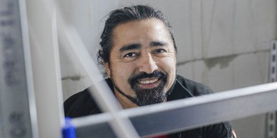 Ein lächelnder Mann mit langen, dunklen Haaren und einem Bart sitzt in einem Raum mit grauen Wänden und einem Metallregal.