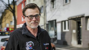 Ein lächelnder Mann mit Brille steht vor einem Gebäude, umgeben von Bäumen und Autos auf einer belebten Straße.