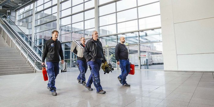 Vier Männer in Arbeitskleidung gehen mit Werkzeugen und Ausrüstung durch eine helle, moderne Halle mit großen Fenstern.