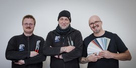 Drei Handwerker stehen lächelnd vor einer neutralen Wand, jeder mit verschiedenen Werkzeugen und Farbmustern in der Hand.