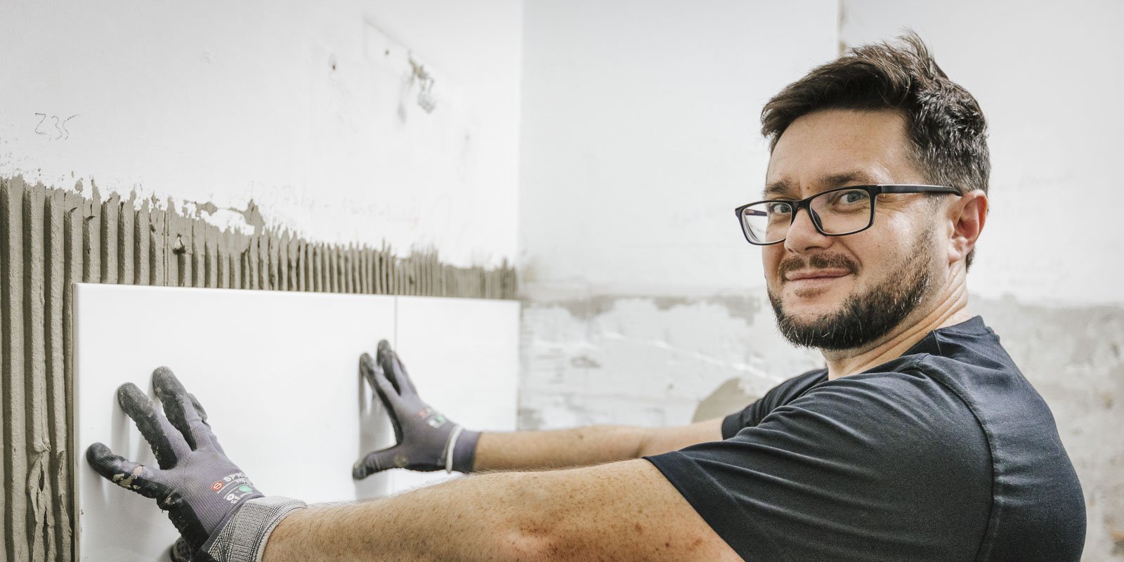 Ein Handwerker mit Brille lächelt, während er eine weiße Fliese an die Wand drückt, umgeben von frischem Mörtel.