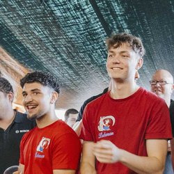 Eine Gruppe junger Männer in roten T-Shirts lächelt und unterhält sich fröhlich unter einem grünen Zelt.