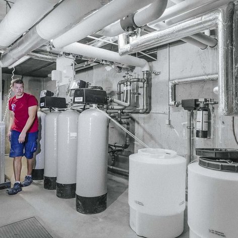 Ein Mann in einem roten T-Shirt steht in einem technischen Raum mit großen Wasserbehältern und Rohren aus Metall.