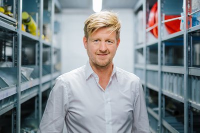 Ein Mann mit blonden Haaren lächelt in einem Lagerraum voller Regale mit Werkzeugen und Ausrüstung.