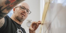 Ein tätowierter Mann mit Brille arbeitet konzentriert an einer Holzoberfläche in einem hellen Raum.