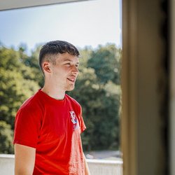 Ein junger Mann mit einem roten T-Shirt lächelt, während er auf einem Balkon steht und die Aussicht genießt.