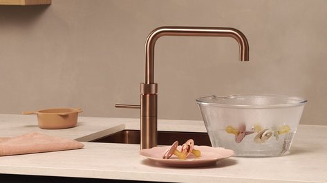 Eine moderne Küchenarmatur aus Kupfer über einer Glasschüssel mit dampfendem Wasser und bunten Schnullern.