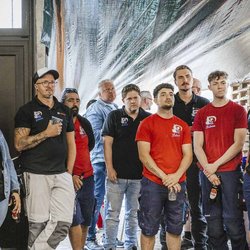 Eine Gruppe von Menschen in Arbeitskleidung hört aufmerksam zu, während sie in einem Raum mit Paletten stehen.