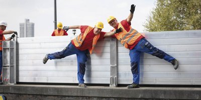 Zwei Bauarbeiter in leuchtenden Westen posieren fröhlich, während sie an einer Wand arbeiten, umgeben von weiteren Kollegen.
