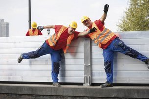 Zwei Bauarbeiter in leuchtenden Westen posieren fröhlich, während sie an einer Wand arbeiten, umgeben von weiteren Kollegen.