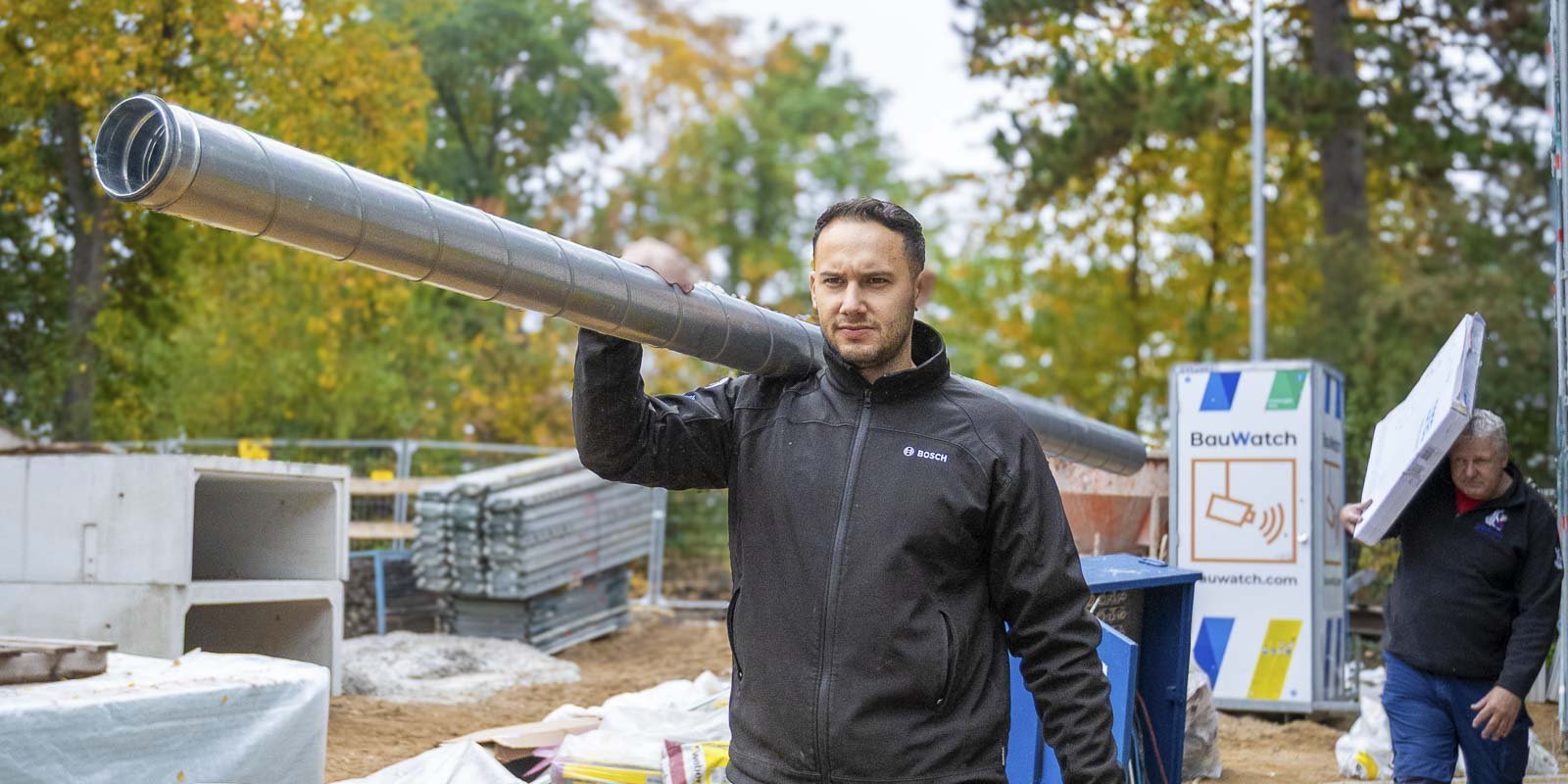 Ein Arbeiter trägt ein langes Metallrohr auf der Schulter, während er auf einer Baustelle mit herbstlichen Bäumen im Hintergrund steht.