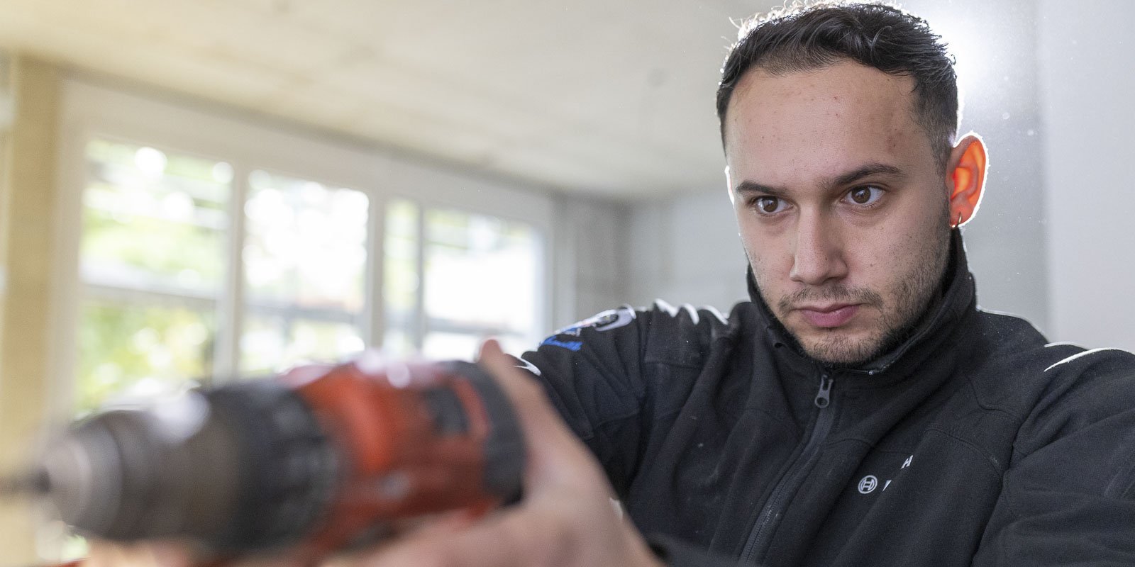 Ein junger Mann mit einem Bohrer in der Hand, konzentriert auf seine Arbeit in einem hellen Raum mit großen Fenstern.