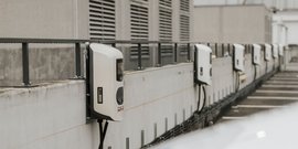 Die Ladestationen für Elektroautos sind entlang einer grauen Wand aufgereiht, umgeben von einem urbanen Umfeld.