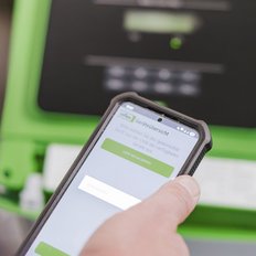 Eine Person hält ein Smartphone, während sie eine App zur Geräteübersicht auf einem grünen Gerät nutzt.