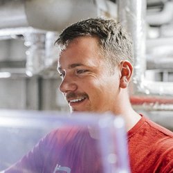 Ein junger Mann mit einem freundlichen Lächeln arbeitet konzentriert in einer industriellen Umgebung mit Rohren im Hintergrund.