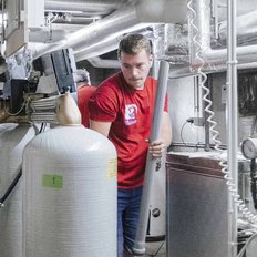 Ein Mann in einem roten T-Shirt arbeitet in einem technischen Raum mit großen Gasflaschen und Rohren.