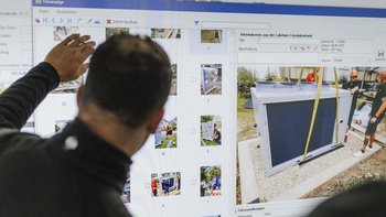 Ein Mann zeigt auf einen Bildschirm mit Bildern von Bauarbeiten und einem großen Gerät, während andere Personen im Hintergrund arbeiten.