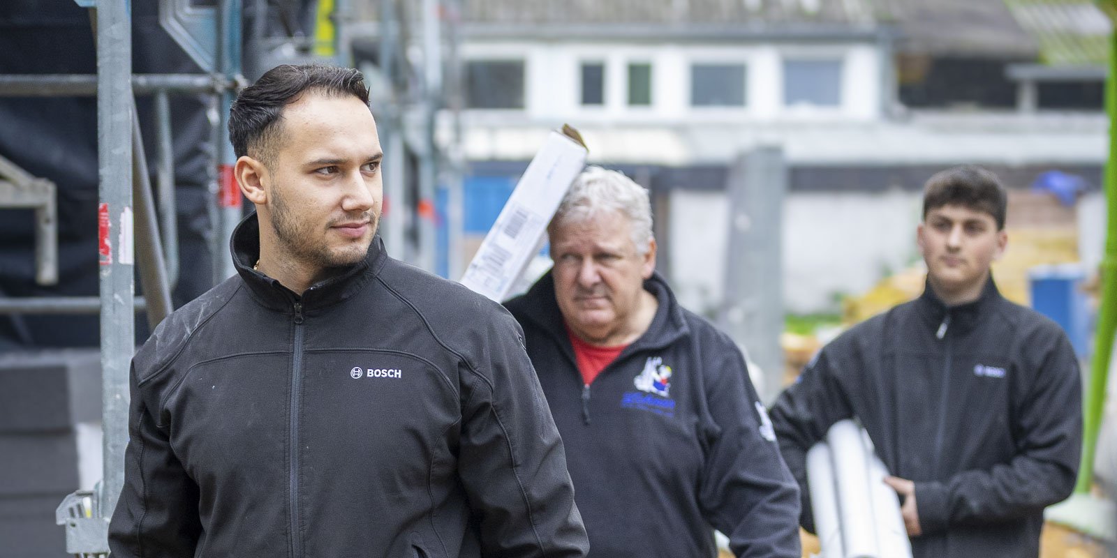 Drei Männer in Arbeitskleidung gehen auf einer Baustelle, während sie Materialien transportieren und konzentriert wirken.