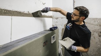 Ein Handwerker verlegt Fliesen an einer Wand, während er sorgfältig Mörtel mit einem Spachtel aufträgt.