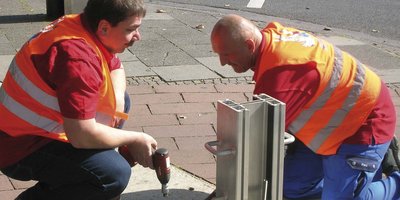 Zwei Arbeiter in leuchtenden Westen arbeiten konzentriert an einem Projekt auf dem Bürgersteig, umgeben von herbstlichen Blättern.