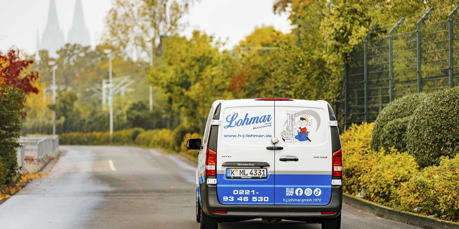Ein Lieferwagen mit dem Logo von Lohmar fährt eine ruhige Straße entlang, umgeben von buntem Herbstlaub.