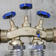 Eine komplexe Rohrleitung mit drei blauen Ventilen, die auf einer grauen Wand montiert ist, zeigt industrielle Details.