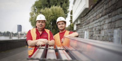 Zwei Bauarbeiter stehen lächelnd vor einer Steinmauer, während sie auf eine Reihe von Metallprofilen zeigen.
