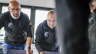 Zwei Männer in dunklen Jacken beugen sich über einen Tisch, während sie konzentriert an einem Projekt arbeiten.