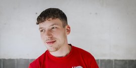 Ein junger Mann mit lockigem Haar trägt ein rotes T-Shirt und lächelt leicht vor einer neutralen Wand.