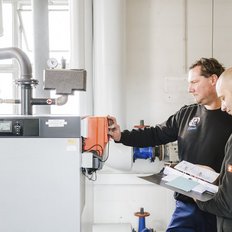 Zwei Techniker überprüfen gemeinsam eine Heizungsanlage, während sie Notizen in einem Ordner machen und Geräte einstellen.