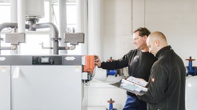 Zwei Techniker überprüfen gemeinsam eine Heizungsanlage, während sie Notizen in einem Ordner machen und Geräte einstellen.