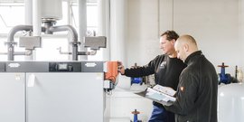 Zwei Techniker überprüfen gemeinsam eine Heizungsanlage, während sie Notizen in einem Ordner machen und Geräte einstellen.