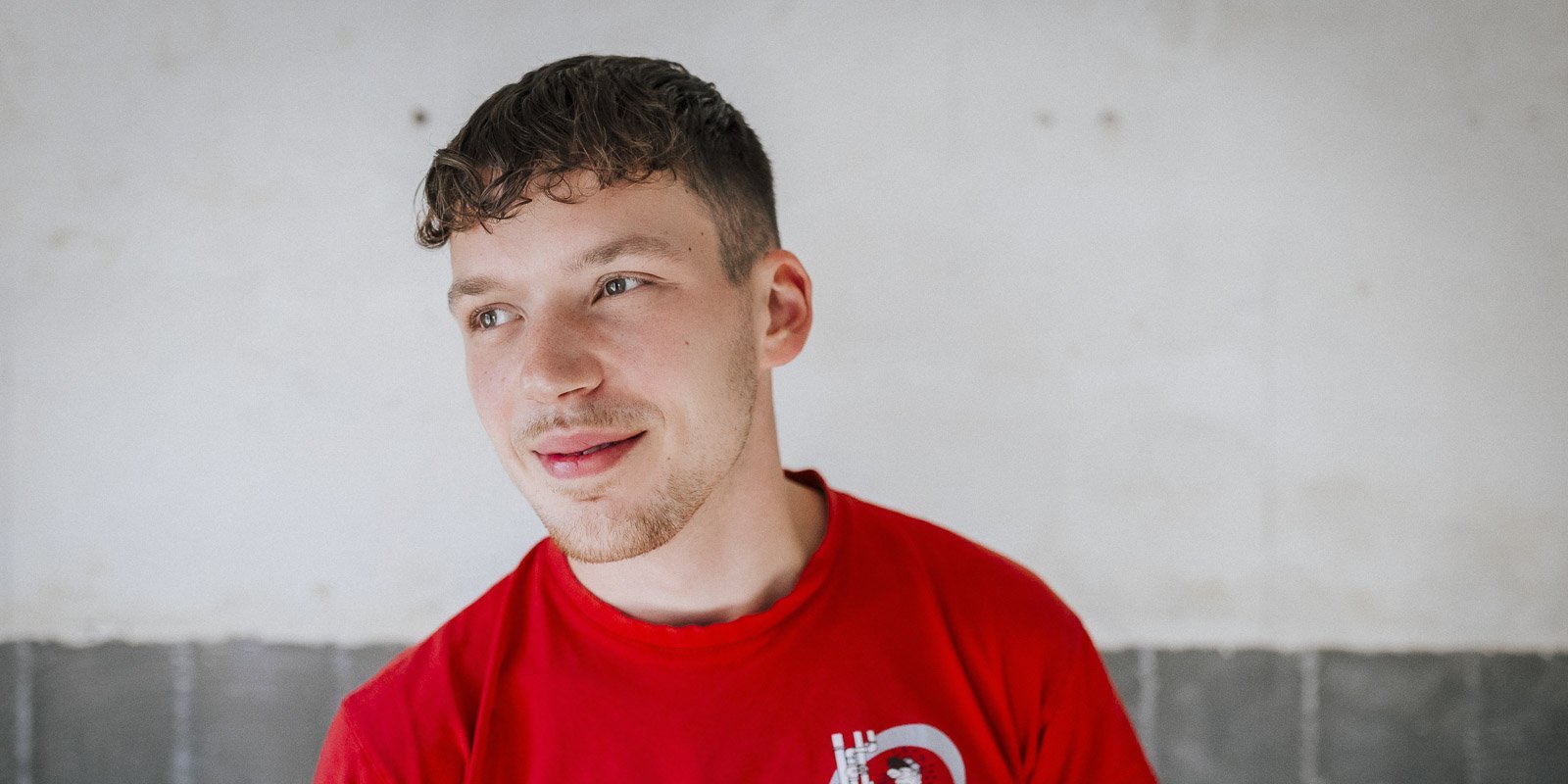 Ein junger Mann mit lockigem Haar trägt ein rotes T-Shirt und lächelt leicht, während er in einem hellen Raum steht.