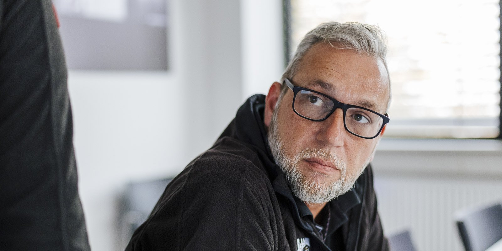 Ein Mann mit Brille und grauem Bart schaut nachdenklich zur Seite, während er in einem modernen Raum sitzt.